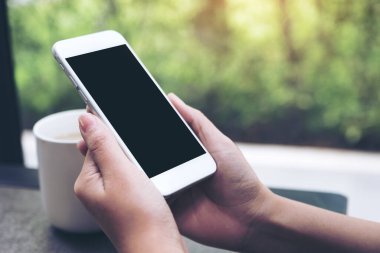 Kahve masasında kahve fincanı olan, boş siyah masaüstü ekranlı, beyaz cep telefonu tutan ellerin görüntüsü.