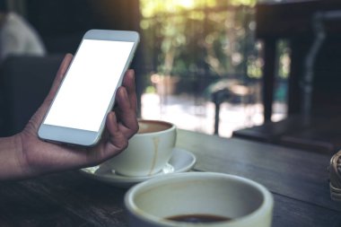 Kafe 'deki ahşap masada kahve fincanlarıyla beyaz cep telefonu ve boş masaüstü ekranlı bir kadının el resmi.
