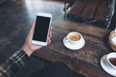 Kafe 'deki ahşap masada kahve fincanlarıyla beyaz cep telefonu ve boş masaüstü ekranlı bir kadının el resmi.