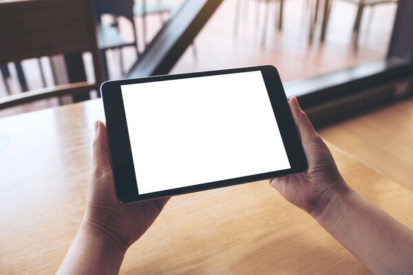 Mockup image of hands holding black tablet pc with blank white desktop screen on wooden table
