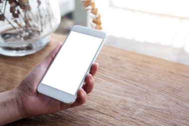Kafedeki ahşap masada boş masaüstü ekranı olan beyaz cep telefonunu tutan el resmi.