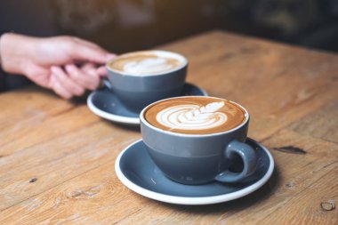 Kafe 'deki eski ahşap masada mavi bir fincan sıcak latte kahve tutan bir elin yakın plan görüntüsü.
