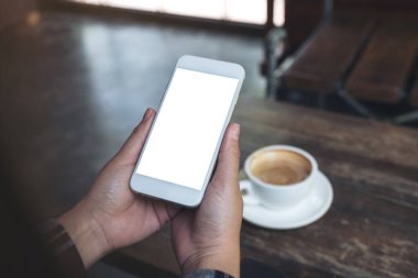 Kafedeki ahşap masada kahve fincanı ve boş masaüstü ekranı olan beyaz cep telefonunu tutan ellerin görüntüsü.
