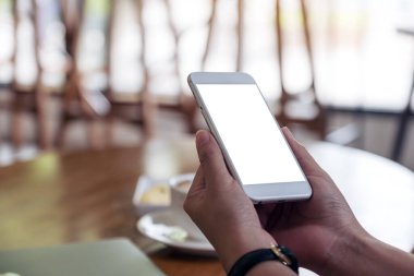 Kafedeki boş masaüstü ekranlı beyaz cep telefonu tutan bir kadının ellerinin görüntüsü.