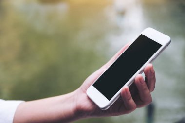 Beyaz akıllı telefonu olan, boş siyah masaüstü ekran ve bulanık yeşil arka planı olan bir kadının görüntüsü.