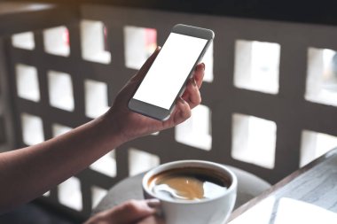 Boş masaüstü ekranı ve kafede kahve fincanı olan beyaz bir cep telefonu tutan bir kadının görüntüsü.