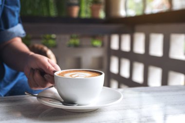 Kafe 'de, elinde bir fincan sıcak kahveyle kalp lattesi tablosu olan bir kadının yakın plan fotoğrafı.