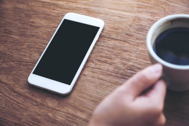 Ahşap masadaki boş masaüstü ekranlı, beyaz cep telefonlu kahve fincanı tutan bir el resmi.