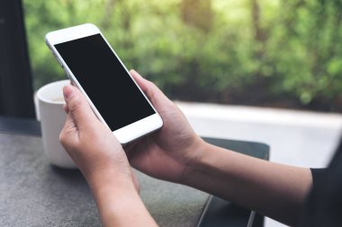 Kahve masasında kahve fincanı olan, boş siyah masaüstü ekranlı, beyaz cep telefonu tutan ellerin görüntüsü.