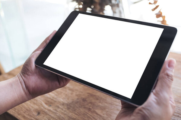 Mockup image of hands holding black tablet pc with blank white desktop screen on wooden table