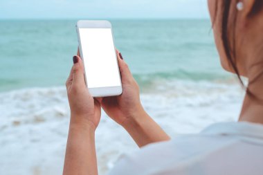 Deniz ve mavi gökyüzü arka planında beyaz cep telefonu ve boş masaüstü ekranı olan bir kadının el görüntüsü.
