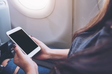 Bulutları ve gökyüzü arkaplanı olan bir uçak penceresinin yanındaki boş siyah masaüstü ekranlı beyaz akıllı telefona bakan bir kadının görüntüsü.