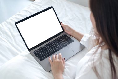 Yatakta oturan Asyalı bir kadının boş beyaz masaüstü klavyesi olan dizüstü bilgisayarı kullanarak ve yazarak görüntüsü. 