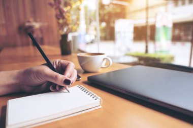Ahşap masa arkasına dizüstü bilgisayar, tablet ve kahve fincanı olan boş bir deftere kadın eli yazarken çekilmiş yakın plan resmi.
