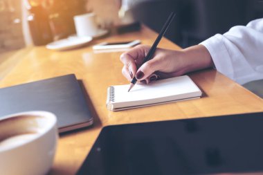 Ahşap masa arkasına dizüstü bilgisayar, tablet ve kahve fincanı olan boş bir deftere kadın eli yazarken çekilmiş yakın plan resmi.