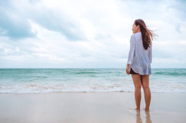 Asyalı bir kadın deniz ve mavi gökyüzü arka planıyla sahilde durup manzarayı seyrediyor.