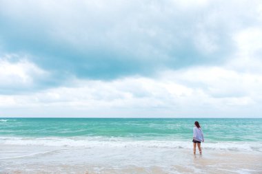 Deniz ve mavi gökyüzü arka planıyla sahilde durup manzaraya bakan bir kadın.