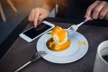 Kafede beyaz tabakta çatalla portakallı pasta keserken ve yerken akıllı telefon kullanan bir kadının yakın plan görüntüsü.