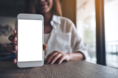 Modern kafedeki boş ekranlı beyaz cep telefonunu tutan ve gösteren bir kadının görüntüsü.
