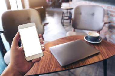 Modern bir kafede beyaz cep telefonu, boş masaüstü ekranı, dizüstü bilgisayarı ve kahve fincanı olan bir adamın ellerinin görüntüsü.