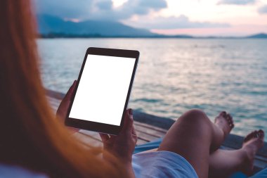 Mavi gökyüzü arka planına sahip deniz kenarında otururken siyah tablet beyaz masaüstü ekran kullanan bir kadının görüntüsü
