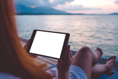 Mavi gökyüzü arka planına sahip deniz kenarında otururken siyah tablet beyaz masaüstü ekran kullanan bir kadının görüntüsü