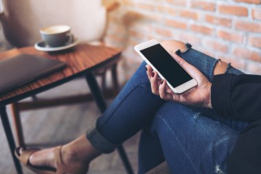 Kafede bacakları çapraz oturan, elinde beyaz cep telefonu ve boş siyah masaüstü ekranı olan bir kadının görüntüsü.