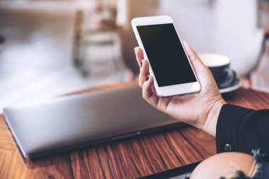 Beyaz cep telefonlu, boş siyah masaüstü ekranlı ve kafedeki ahşap masadaki dizüstü bilgisayarlı bir kadının el resmi.