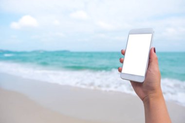 Deniz ve mavi gökyüzü arkaplanı önünde boş masaüstü ekranı olan beyaz cep telefonu tutan bir kadının el görüntüsü.