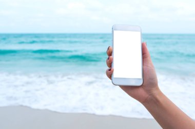 Deniz ve mavi gökyüzü arkaplanı önünde boş masaüstü ekranı olan beyaz cep telefonu tutan bir kadının el görüntüsü.