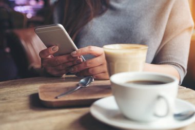 Kafedeki ahşap masada elinde kahve fincanları olan akıllı bir telefonu tutan ve kullanan bir kadının yakın plan görüntüsü.