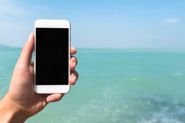 Bir el ele tutuşmanın ve açık siyah masaüstü ekranlı beyaz cep telefonunu deniz ve mavi gökyüzü arkaplanının önünde göstermenin görüntüsü.