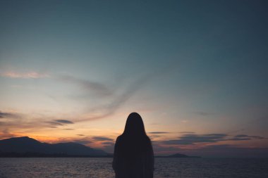 Sahilde deniz ve gün batımına bakan bir kadının siluetleri.