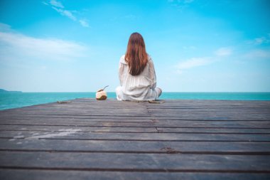 Beyaz elbiseli güzel bir Asyalı kadın oturmuş denize bakıyor ve mavi gökyüzünü ahşap balkonda rahatlamış hissediyor.