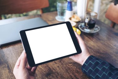 Boş masaüstü beyaz ekranlı siyah tableti ve kafenin arka planındaki ahşap masada kahve fincanını tutan ellerin görüntüsü.
