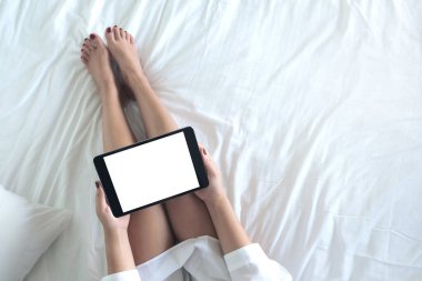 Beyaz bir yatakta otururken elinde boş masaüstü beyaz ekranı olan siyah tablet bir kadının en üstteki görünüm görüntüsü