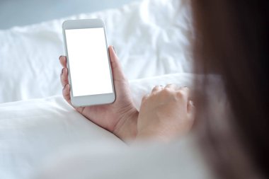 Beyaz yatakta oturan, boş masaüstü ekranlı, beyaz cep telefonlu bir kadının el resmi.