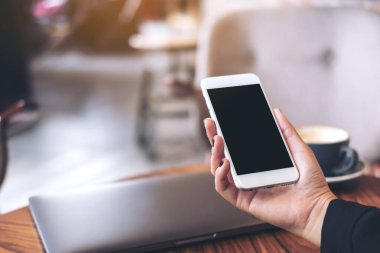 Beyaz cep telefonlu, boş siyah masaüstü ekranlı ve kafedeki ahşap masadaki dizüstü bilgisayarlı bir kadının el resmi.