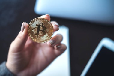 Ofisinde dizüstü bilgisayarı ve cep telefonu olan altın renkli bir Bitcoin tutan bir iş kadını resmi.