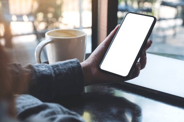 Kafedeki masada kahve fincanı ve dizüstü bilgisayarı olan, boş beyaz ekranlı, siyah cep telefonlu bir kadının el resmi.