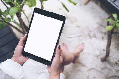 Kum ve plaj arka planına sahip boş beyaz masaüstü ekranlı siyah tableti tutan bir kadının el resmi.