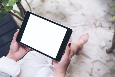 Kum ve plaj arka planına sahip boş beyaz masaüstü ekranlı siyah tableti tutan bir kadının el resmi.