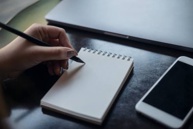 Masanın üzerinde cep telefonu ve dizüstü bilgisayarı olan beyaz bir deftere yazı yazan bir kadının ellerinin yakın plan resmi. 