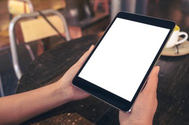 Siyah tableti beyaz masaüstü ekranı ve kafedeki ahşap masada kahve fincanı ile tutan ellerin görüntüsü. 