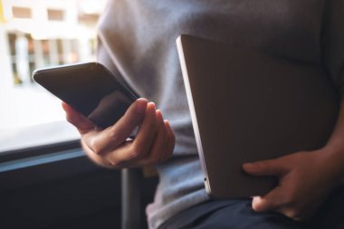 Bilgisayarı elinde tutarken siyah akıllı bir telefonu kullanan ve ona bakan bir kadının yakın plan fotoğrafı.