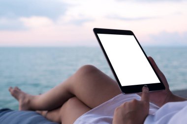 Mavi gökyüzü arkaplanlı deniz kenarında otururken elinde beyaz ekranlı siyah tablet kullanan bir kadının görüntüsü.