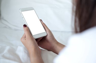 Boş masaüstü ekranlı beyaz cep telefonu tutan bir kadının görüntüsü beyaz yatakta oturuyor.