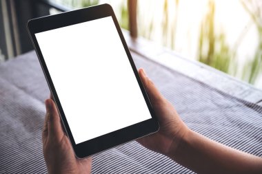 Kafede bulanık arkaplanı olan boş beyaz masaüstü ekranlı siyah tableti tutan ellerin görüntüsü 