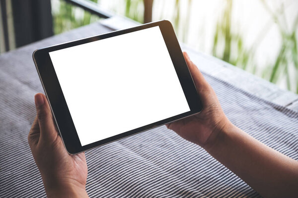 Mockup image of hands holding black tablet pc with blank white desktop screen on table with blur background in cafe 