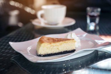 Bir parça peynirli kek ve beyaz tabakta krem şanti. Kafede kahve fincanı var.